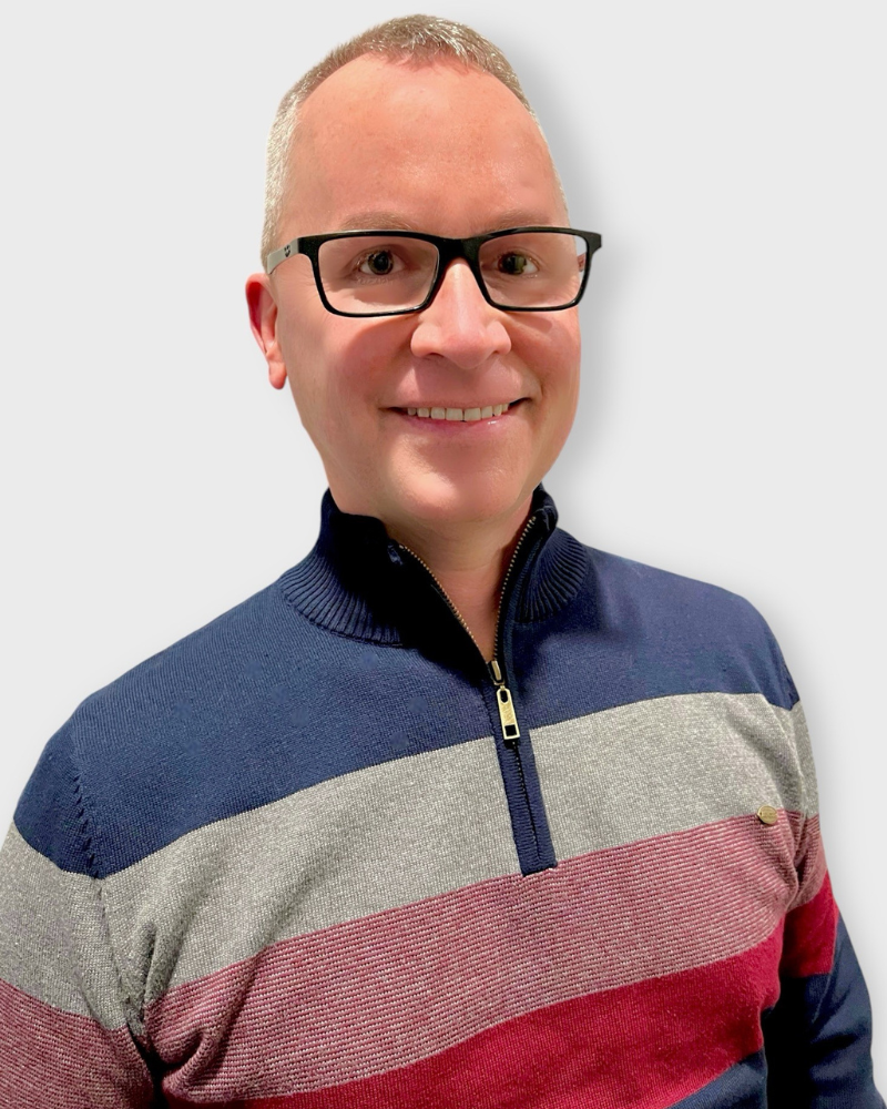 Portrait of a smiling man with short hair and glasses wearing a color-block quarter-zip sweater, photographed against a light background.